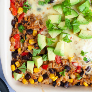 A ligth brown ceramic dish with colorful ingredients like black beans, corn, rice, melted cheese, bell pepper strips, chopped avocado, chopped green herbs.