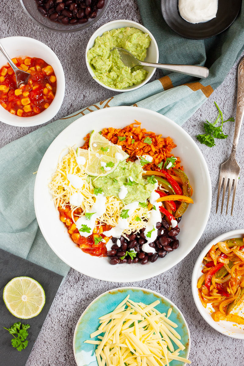 A large white plate in the middle with colorful ingredients like black beans, corn, shredded cheese, bell pepper strips, lime, orange rice. Small white bowl are scattered around with the same ingredients.