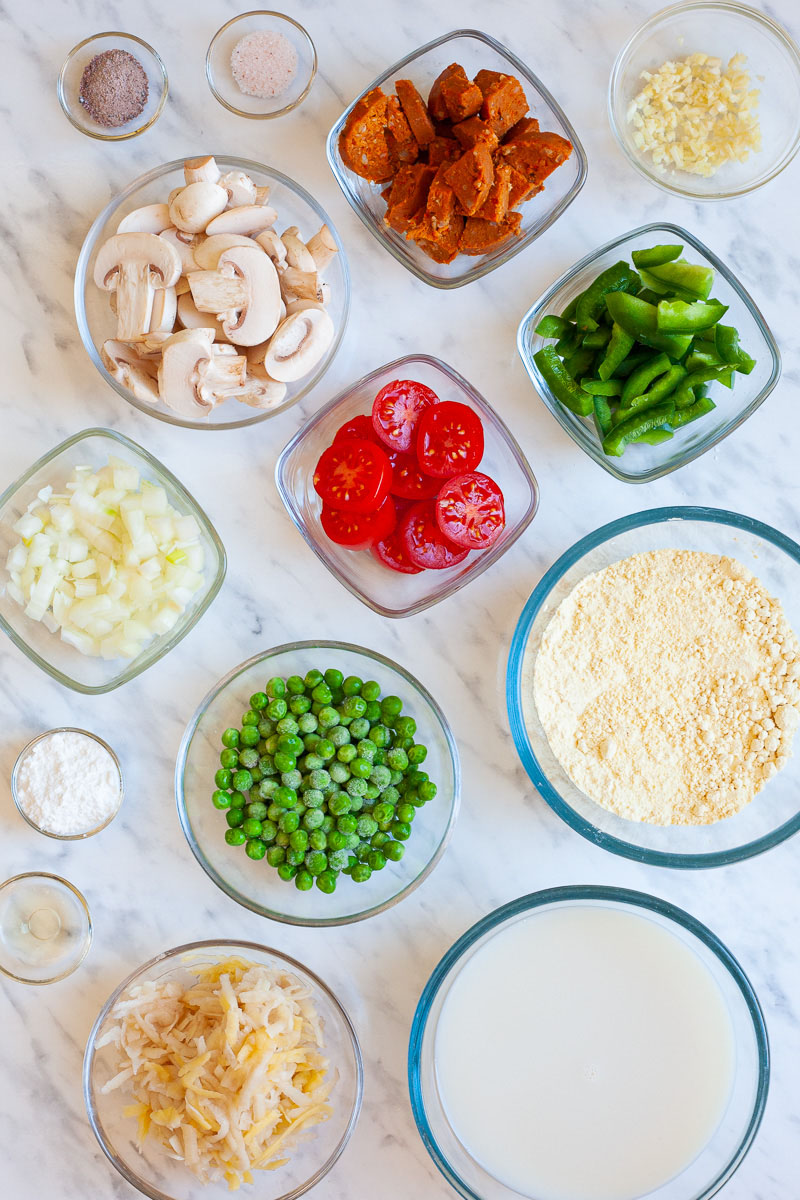 The ingredients of a breakfast casserole is small glass bowls: shredded potato, green peas, chopped onion, chopped garlic, cherry tomatoes, sausage slices, mushroom slices, bell pepper slices, milk, chickpea flour, baking powder, black salt, himalaya salt and apple cider vinegar