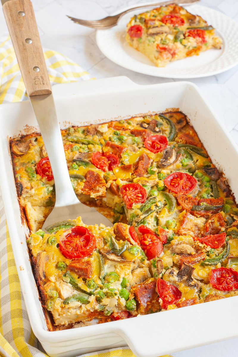 Cherry tomatoes, mushroom slices, bell pepper slices on top of a yellow cake-like dough is in a white casserole dish. A slice served on a white plate is in the background