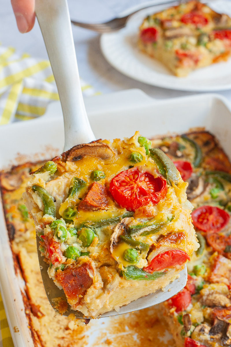 A slice of breakfast casserole with cherry tomatoes, mushroom slices, bell pepper slices on top of a yellow cake-like dough ilifted up with a slotted turner. The white casserole dish is in the background