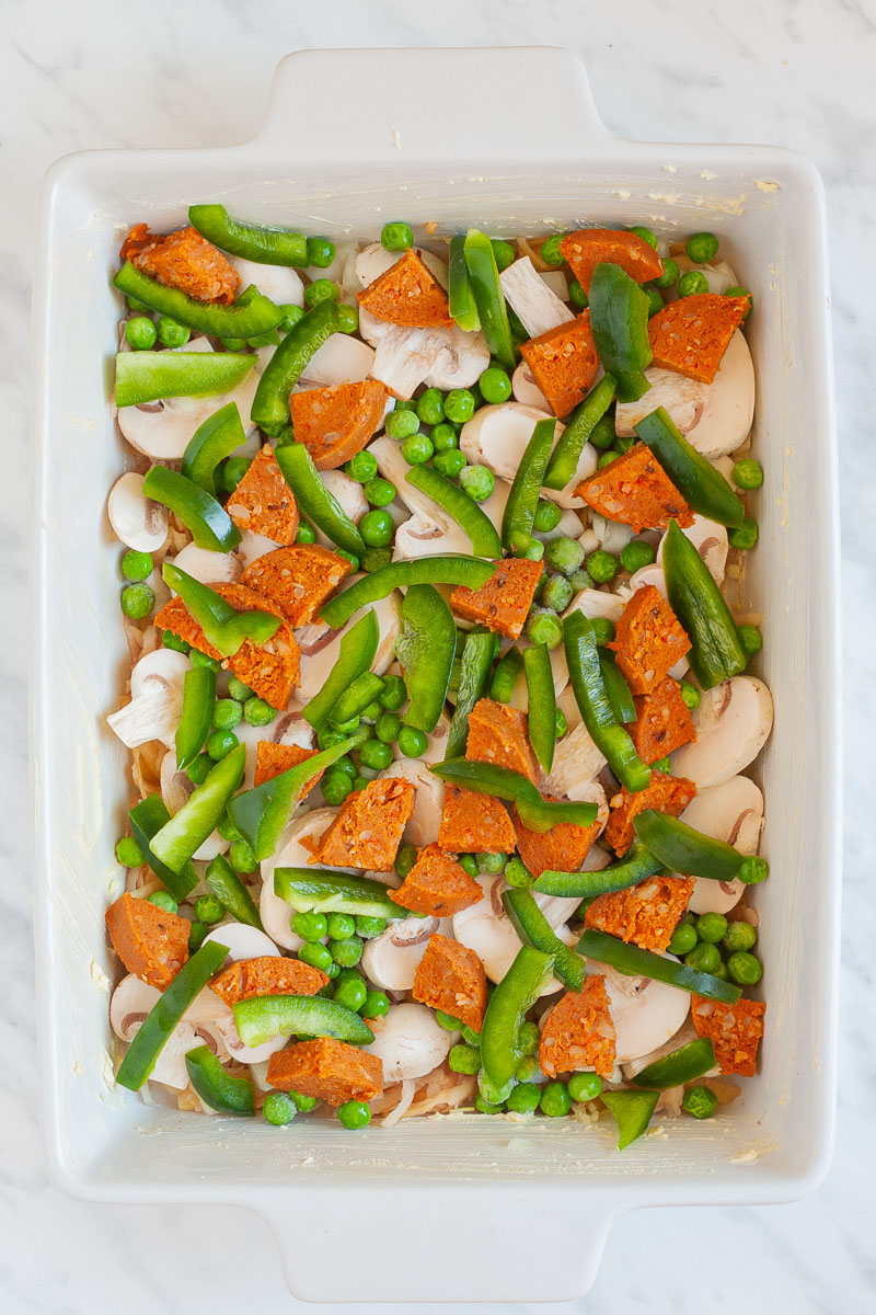 A white casserole dish from above from filled with cherry tomato halves, bell pepper slices, sausage slices, green peas, mushroom slices