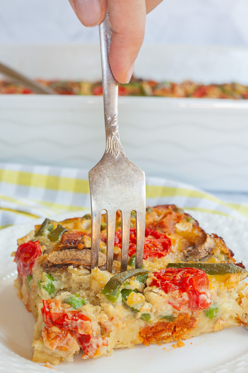 A slice of breakfast casserole with cherry tomatoes, mushroom slices, bell pepper slices on top of a yellow cake-like dough is served on a white plate with a fork piercing in the middle. The white casserole dish is in the background