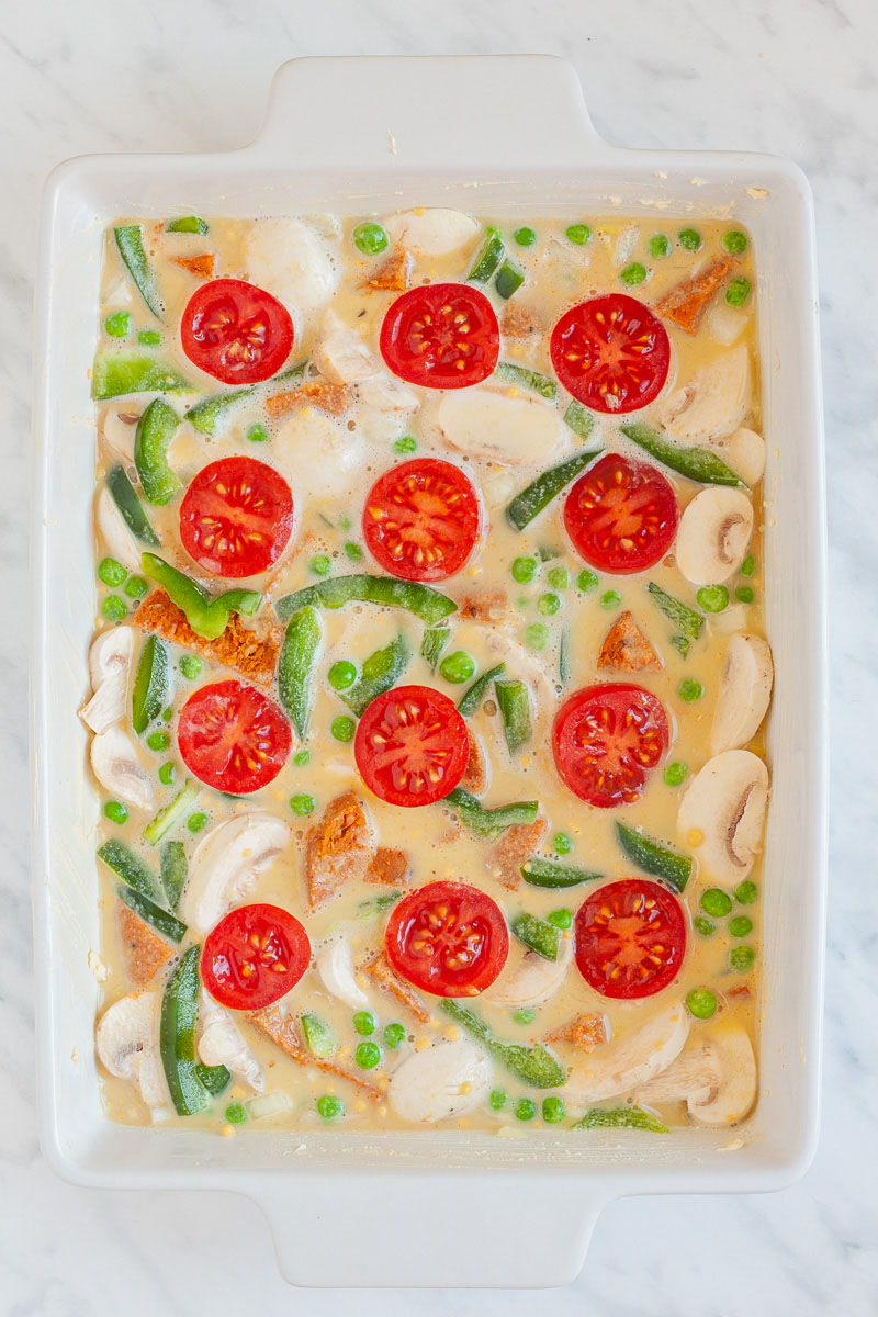 A white casserole dish from above from filled with cherry tomato halves, bell pepper slices, green peas, mushroom slices in a yellow liquid