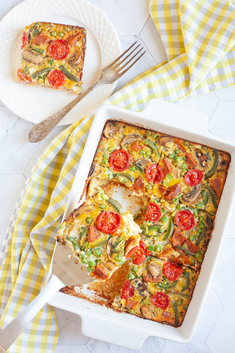 A slice of breakfast casserole with cherry tomatoes, mushroom slices, bell pepper slices on top of a yellow cake-like dough is served on a white plate with a fork. The white casserole dish is in the middle with a slotted turner taking a piece.