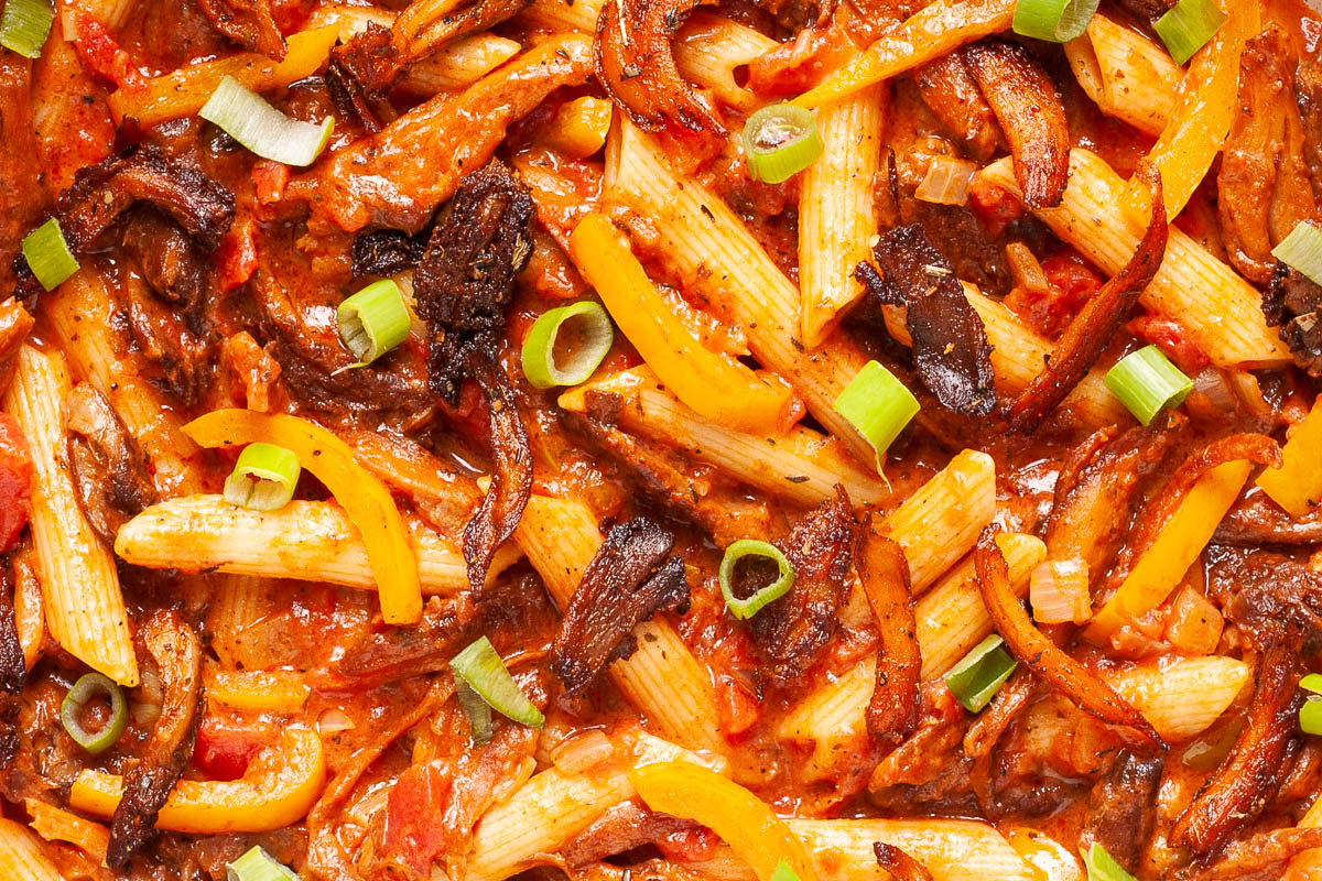 Penne pasta with yellow bell pepper slices, brown mushroom shreds and spring onion rings in a creamy red sauce from above.