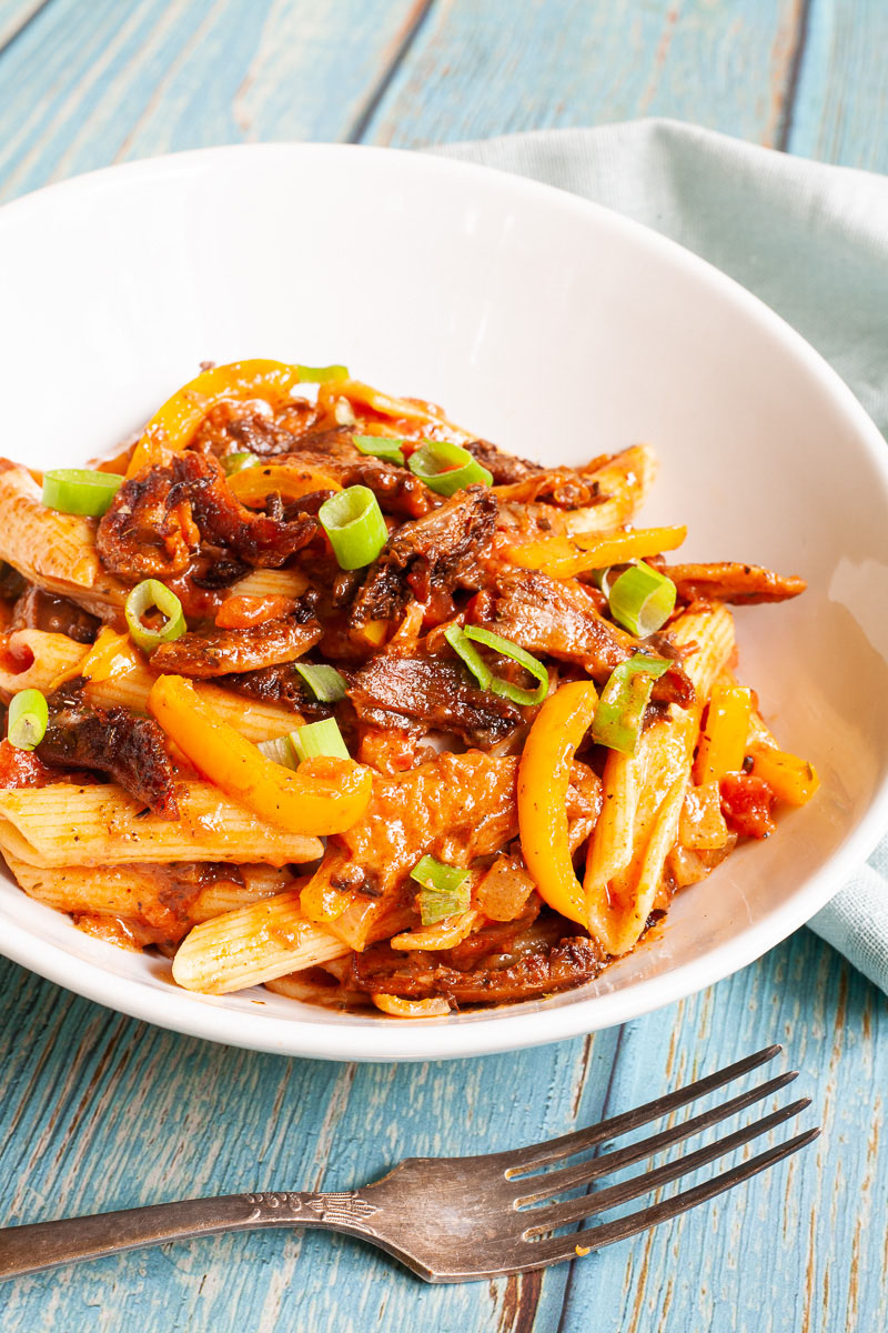 Penne pasta with yellow bell pepper slices, brown mushroom shreds and spring onion rings in a creamy red sauce served in a white bowl. A fork is placing next to the bowl.