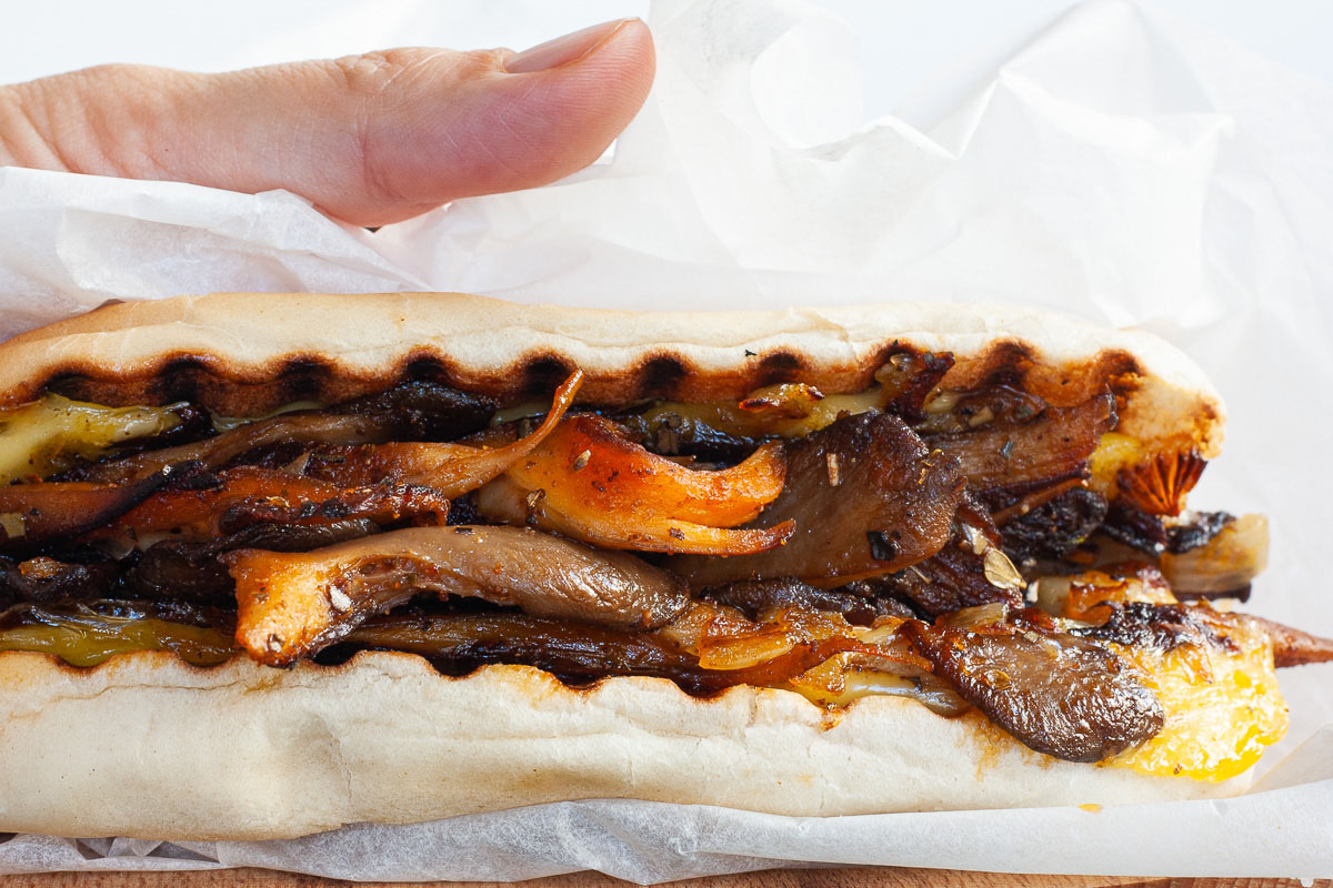A hand is holding a closed bread loaf stuffed with brown mushroom shreds and melted cheese wrapped in a white paper