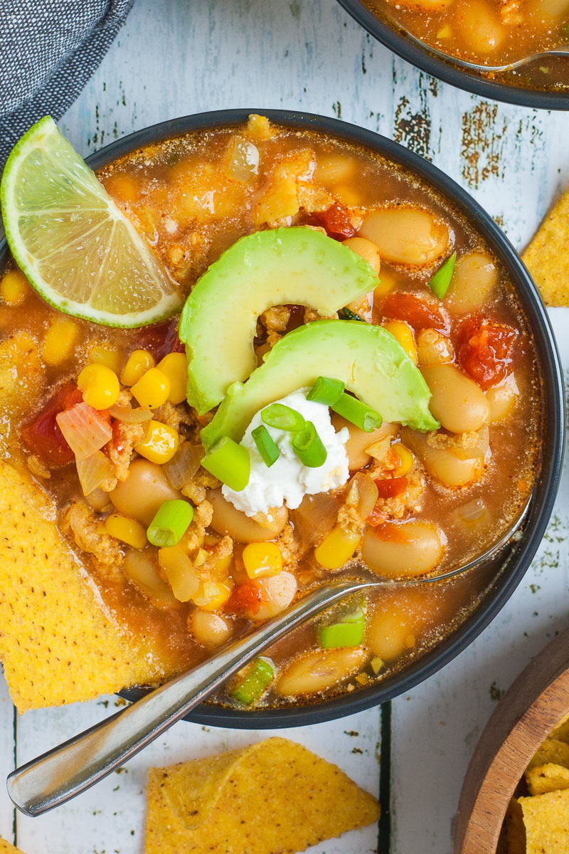 1 black bowl with orange liquid, beans, corn, chopped tomaotes, mince, avocado slices, chopped spring onion, lime slices, and tortilla chips. More tortilla chips are scattered around