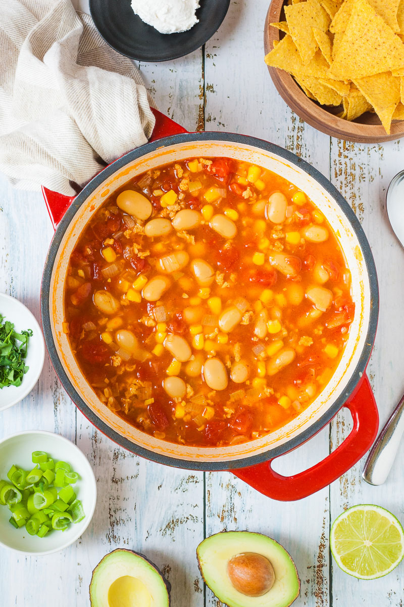 A red Dutch oven is from above with orange soup, white beans, corn, chopped tomato, mince. Half avocado, lime and small bowls of chopped pcilantro and spring onion is around with some tortilla chips and a bit of sour cream on a black plate.