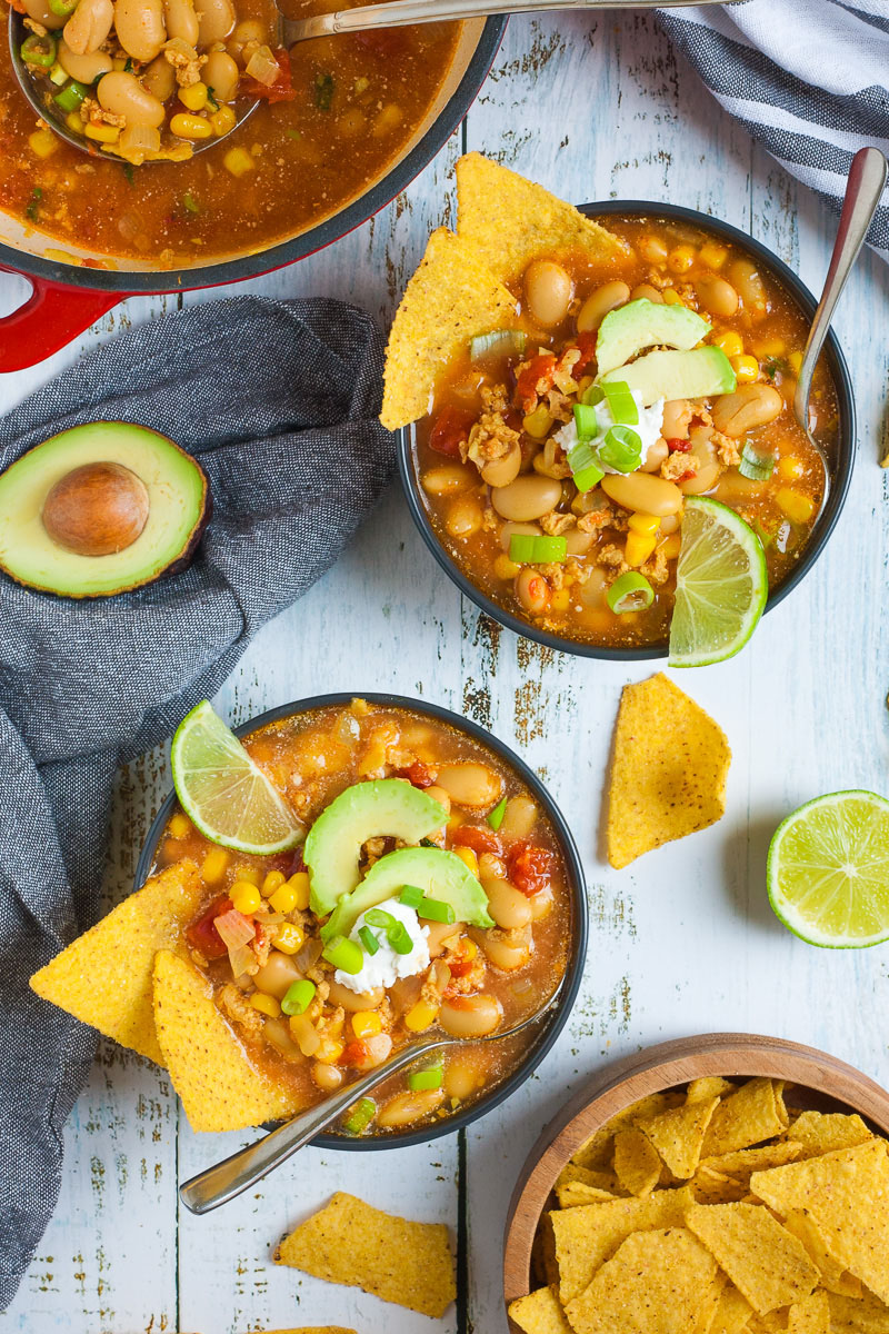 2 black bowls with orange liquid, beans, corn, chopped tomaotes, mince, avocado slices, chopped spring onion, lime slices, and tortilla chips. The red Dutch oven with more soup, half of avocado and more tortilla chips are scattered around
