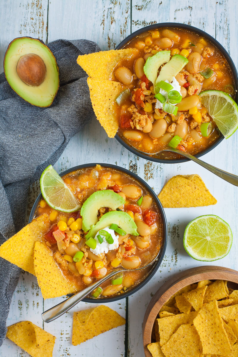 2 black bowls with orange liquid, beans, corn, chopped tomaotes, mince, avocado slices, chopped spring onion, lime slices, and tortilla chips. Half of avocado and more tortilla chips are scattered around