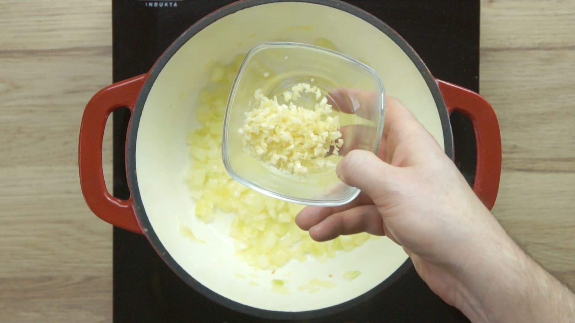 A hand is holding a bowl of chopped garlic to be added to chopped onions in a red enameled Dutch oven. 