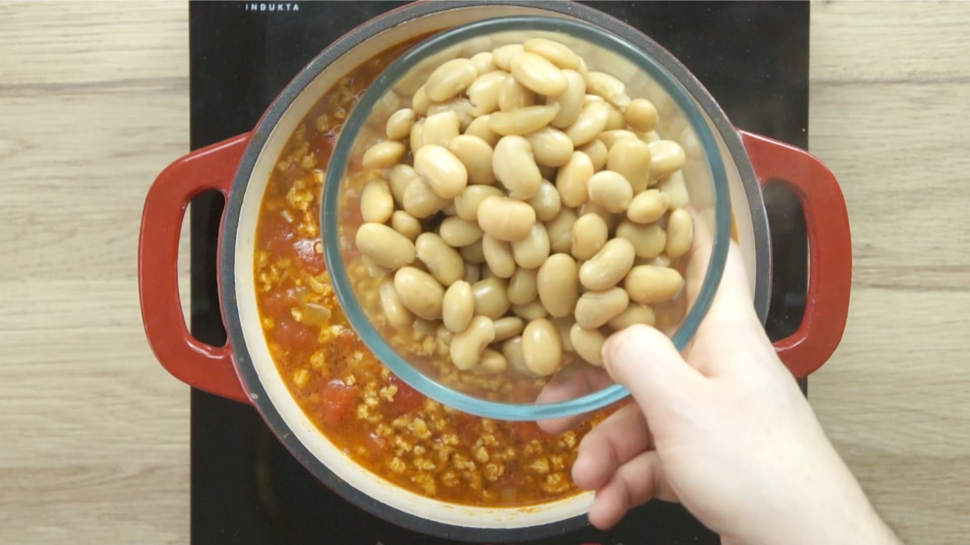 A hand is holding a bowl of chopped white beans to be added to a tomato colored soup below.