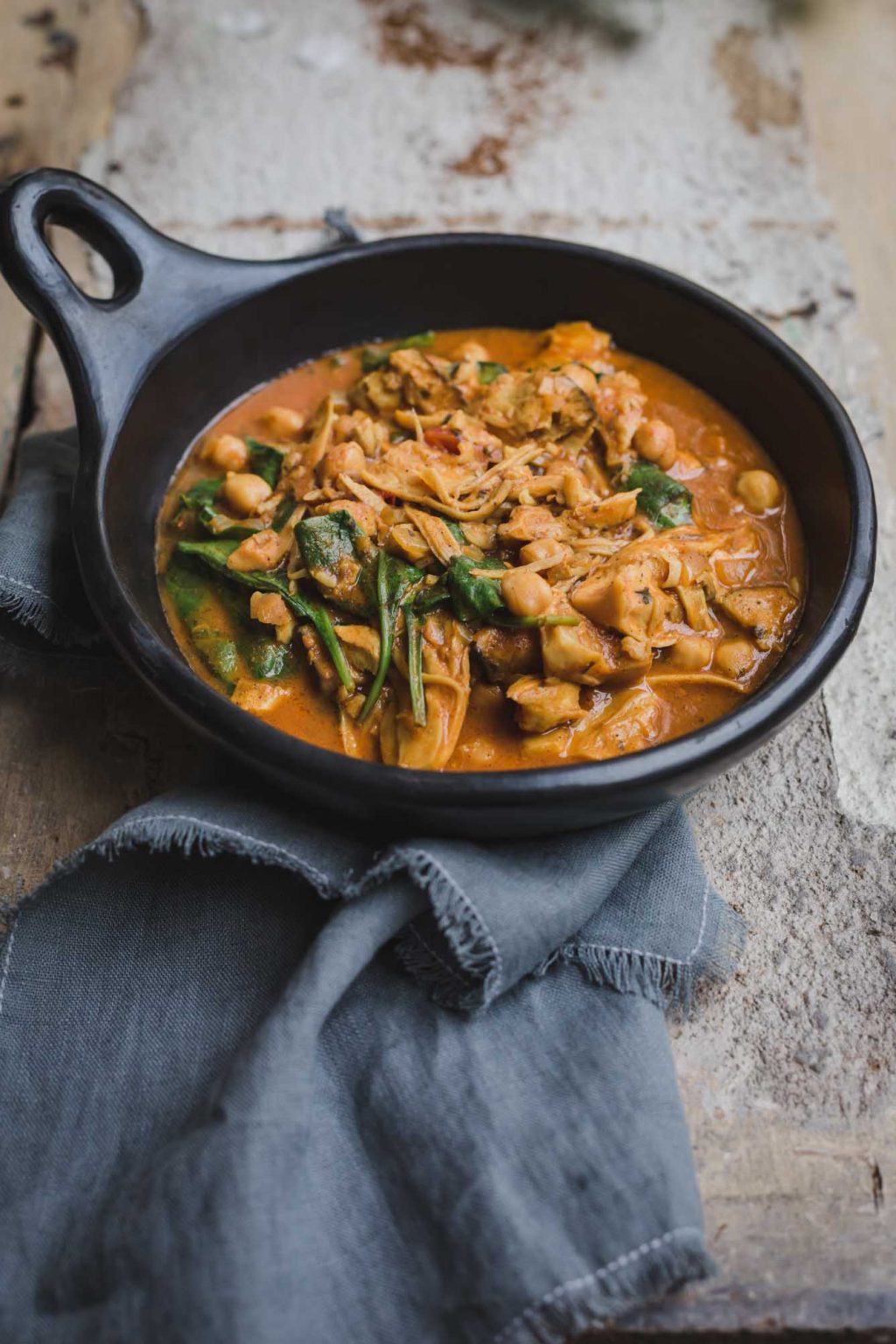 A dark brown sauce is served in a shallow black bowl with shredded mushroom, chickpeas and wilted spinach leaves