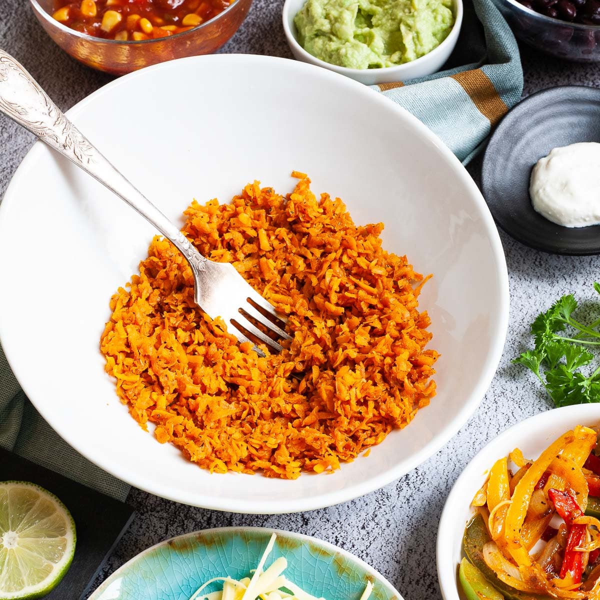 A large white plate in the middle orange riced sweet potato. Small white bowl are scattered around with colorful ingredients like black beans, corn, shredded cheese, bell pepper strips, lime, orange rice.