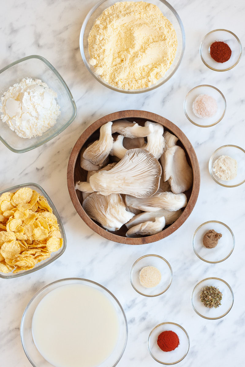 Lots of bowls with the ingredients to make vegan fried chicken: oyster mushrooms, different spices, yellow flour, starch, milk, corn flakes