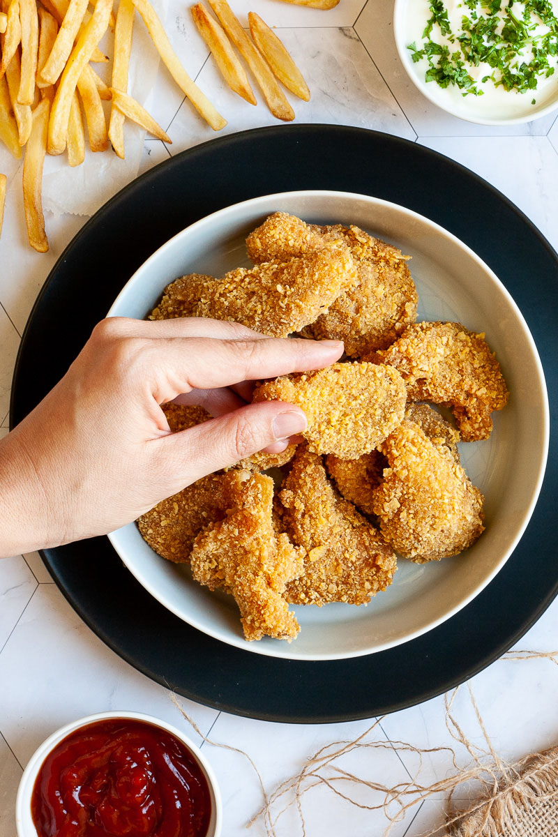 Brown breaded nuggets in a round plate. A hand is holindg one piece. French fries to the side with two small bowls. One with ketchup and the other is a white sauce with chopped green herbs.