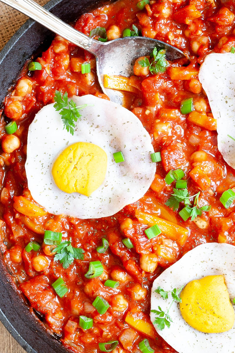 A cast iron skillet from above. 3 fried egg lookalikes with white and yolk part on top of a red chunky sauce sprinkled with green spring onion rings and chopped parsley. You can see chickpeas, chopped tomato and chopped yellow bell peppers.