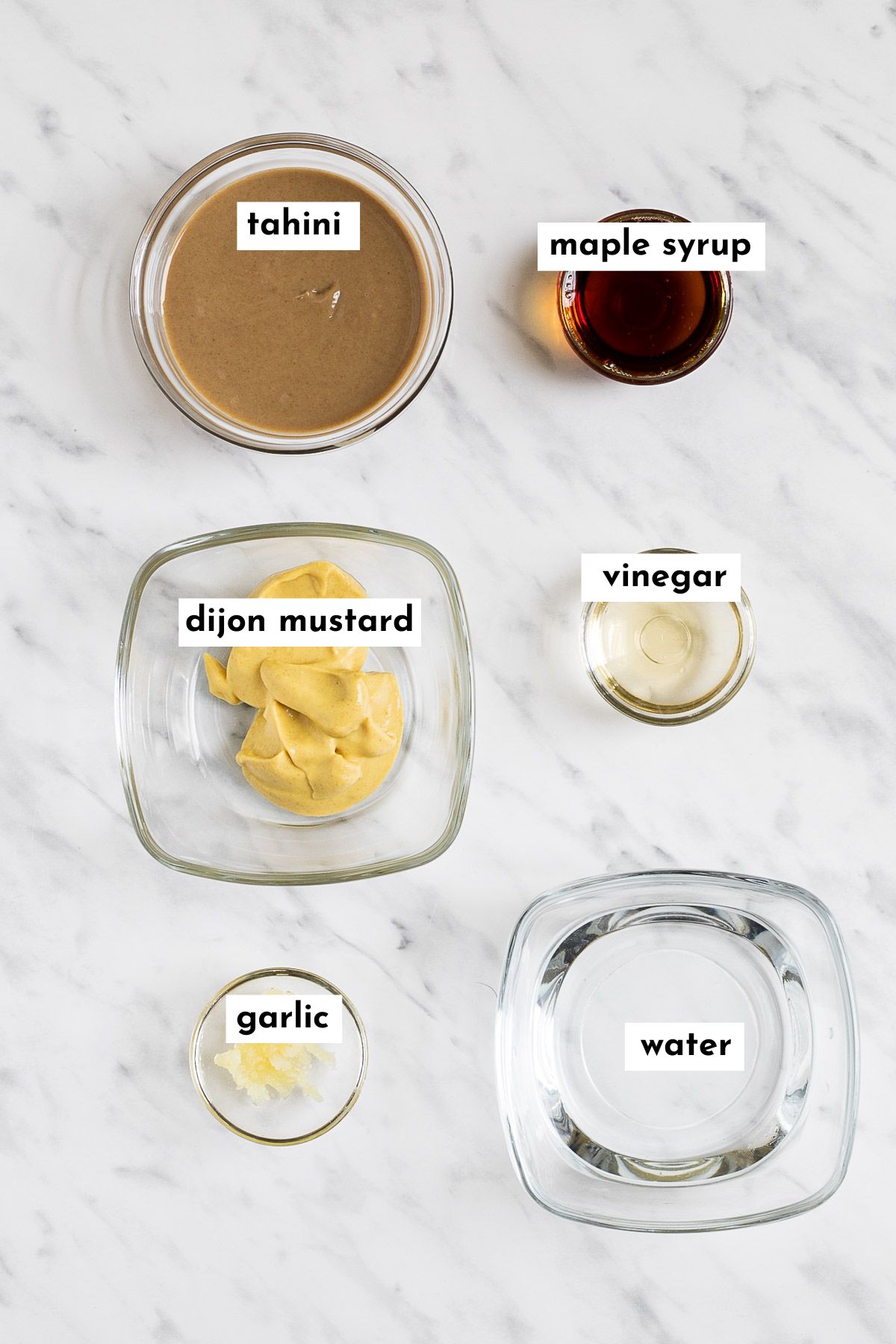 The ingredients of maple dijon tahini dressing is in small glass bowls. These are light brown tahini, mustard, maple syrup, water, vinegar and minced garlic. 
