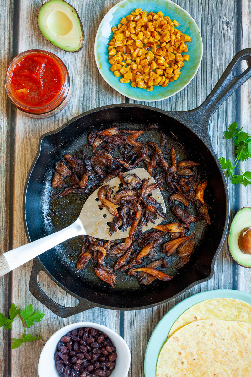 Crispy dark brown oyster mushroom shreds in a black cast-iron skillet. A spatula is holding up a handful. One small bowl with black beans, one with red sauce and one blue plate with corn
