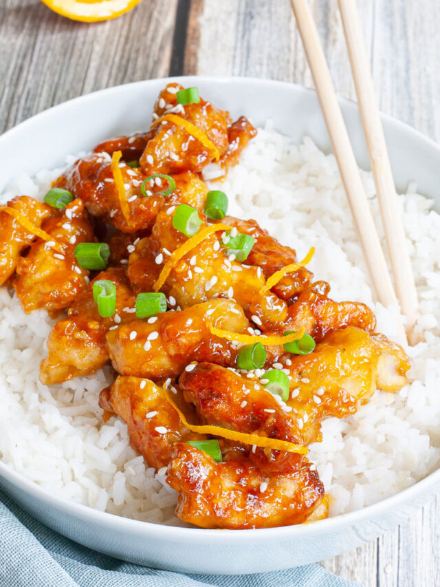 A large white bowl with white rice and sticky brown battered strips on top sprinkled with sesame seeds, orange peels and chopped spring onion