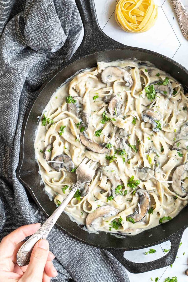 Cast iron skillet with tagliatelle in a creamy light brown sauce with sliced mushrooms and freshly chopped green herbs. A hand is holding a fork to the middle.