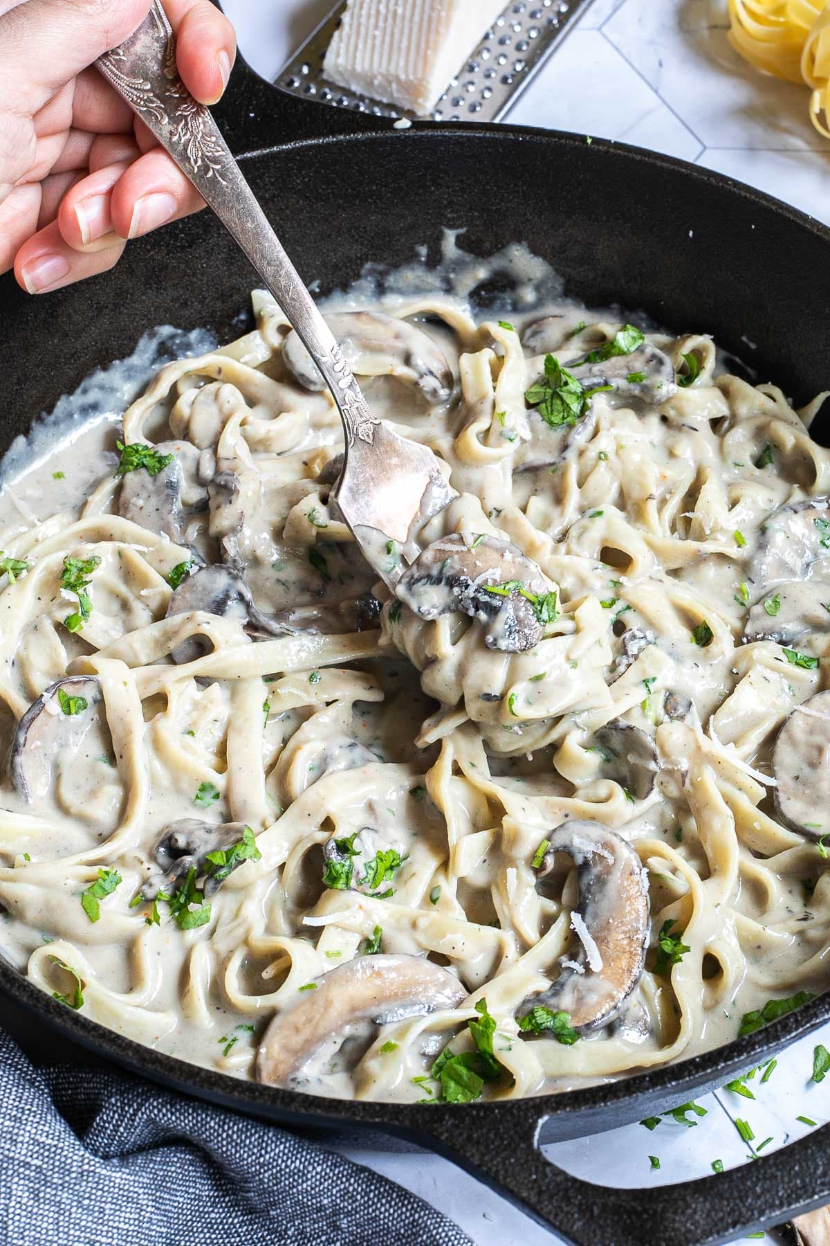 Cast iron skillet with tagliatelle in a creamy light brown sauce with sliced mushrooms and freshly chopped green herbs. A hand is holding a fork to the middle.
