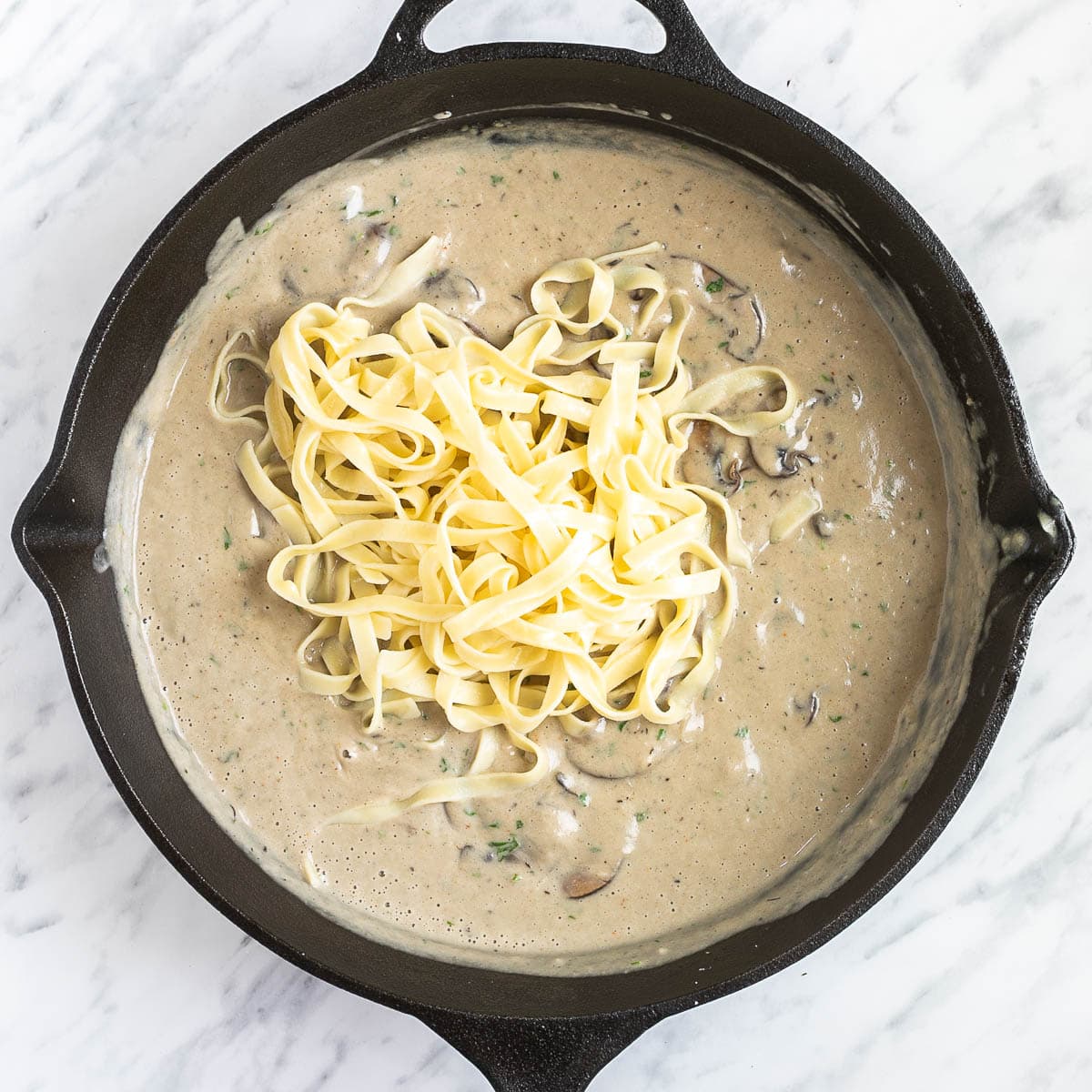 Cast iron skillet with a smooth light brown sauce, mushroom and cooked tagliatelle pasta.