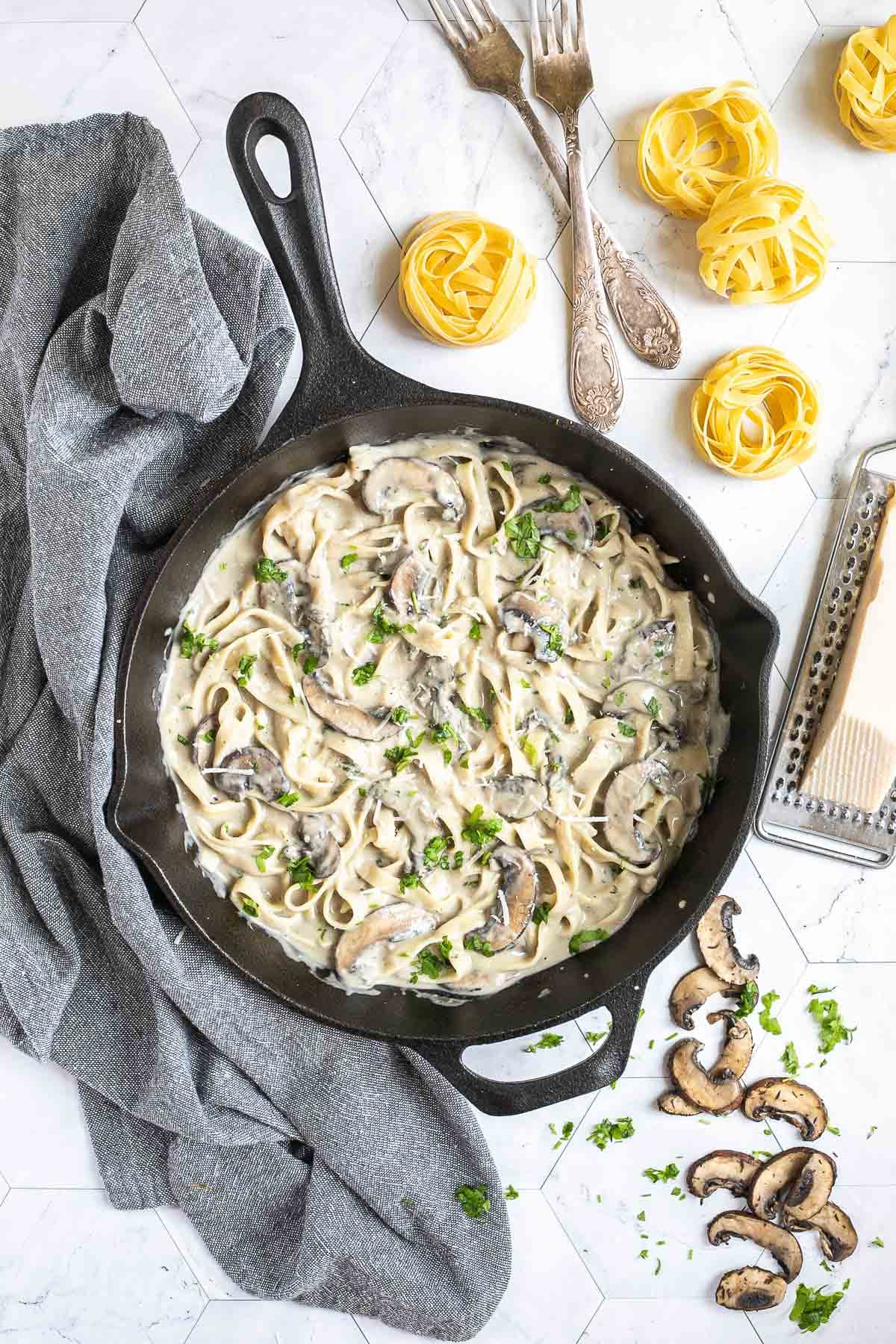 Cast iron skillet with tagliatelle in a creamy light brown sauce with sliced mushrooms and freshly chopped green herbs. Dry tagliatelle and a cheese grater with a stick of parmesan is next to it.