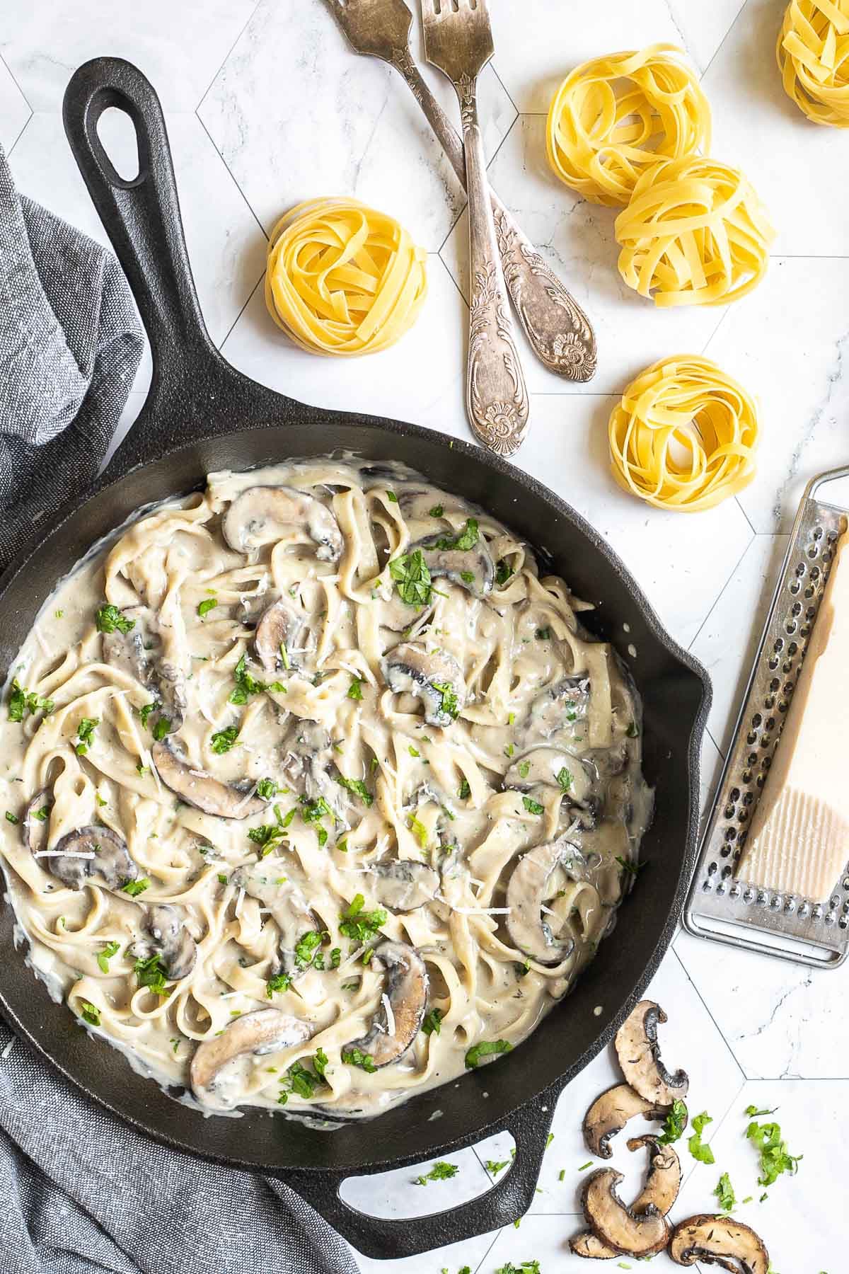 Cast iron skillet with tagliatelle in a creamy light brown sauce with sliced mushrooms and freshly chopped green herbs. Dry tagliatelle and a cheese grater with a stick of parmesan is next to it.