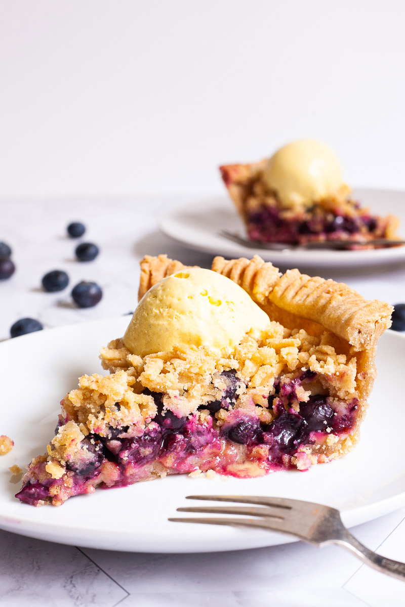 One slice of blueberry pie on a white plate with vibrant purple berries, crumble topping and a scoop of yellow ice cream. Another plate with a slice is in the background. Fresh blueberries are scattered around.