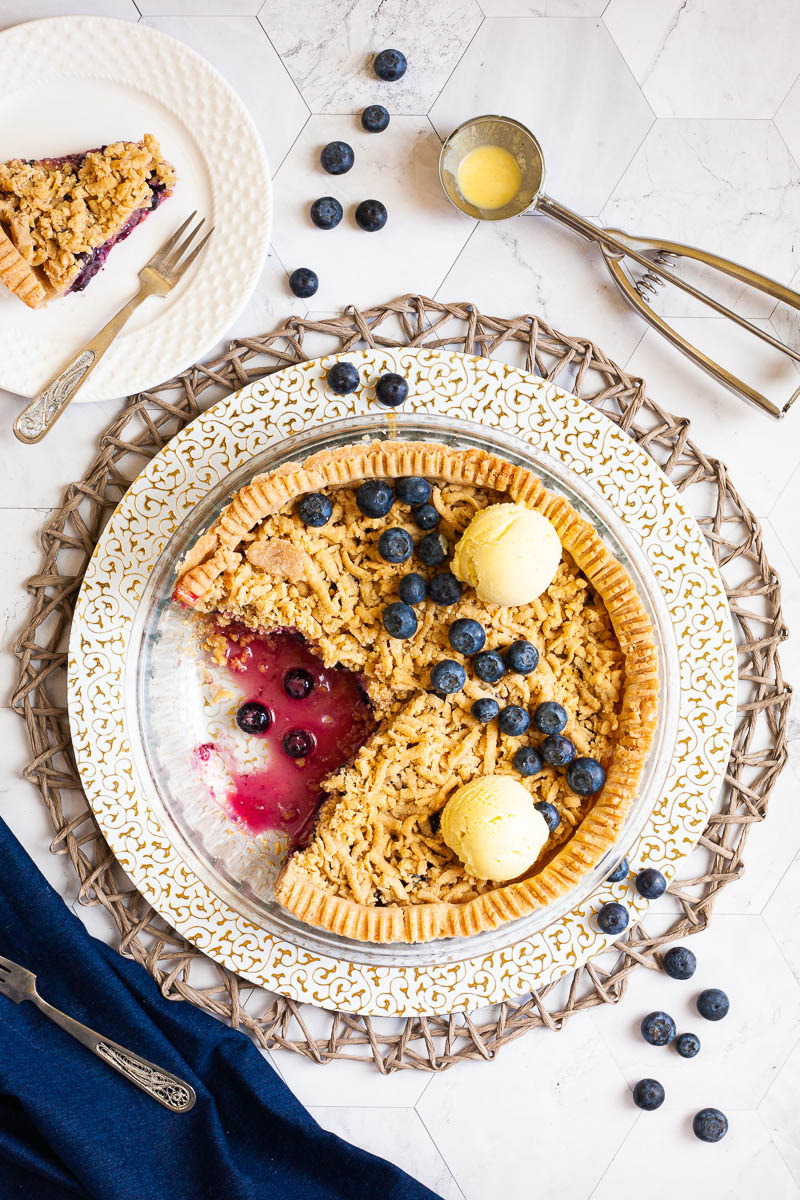 A whole blueberry pie from above with fresh blueberries on top as decoration, crumble topping and 2 scoops of yellow ice cream. 2 slices are missing. A plate with one slice is in the background. Fresh blueberries are scattered around.