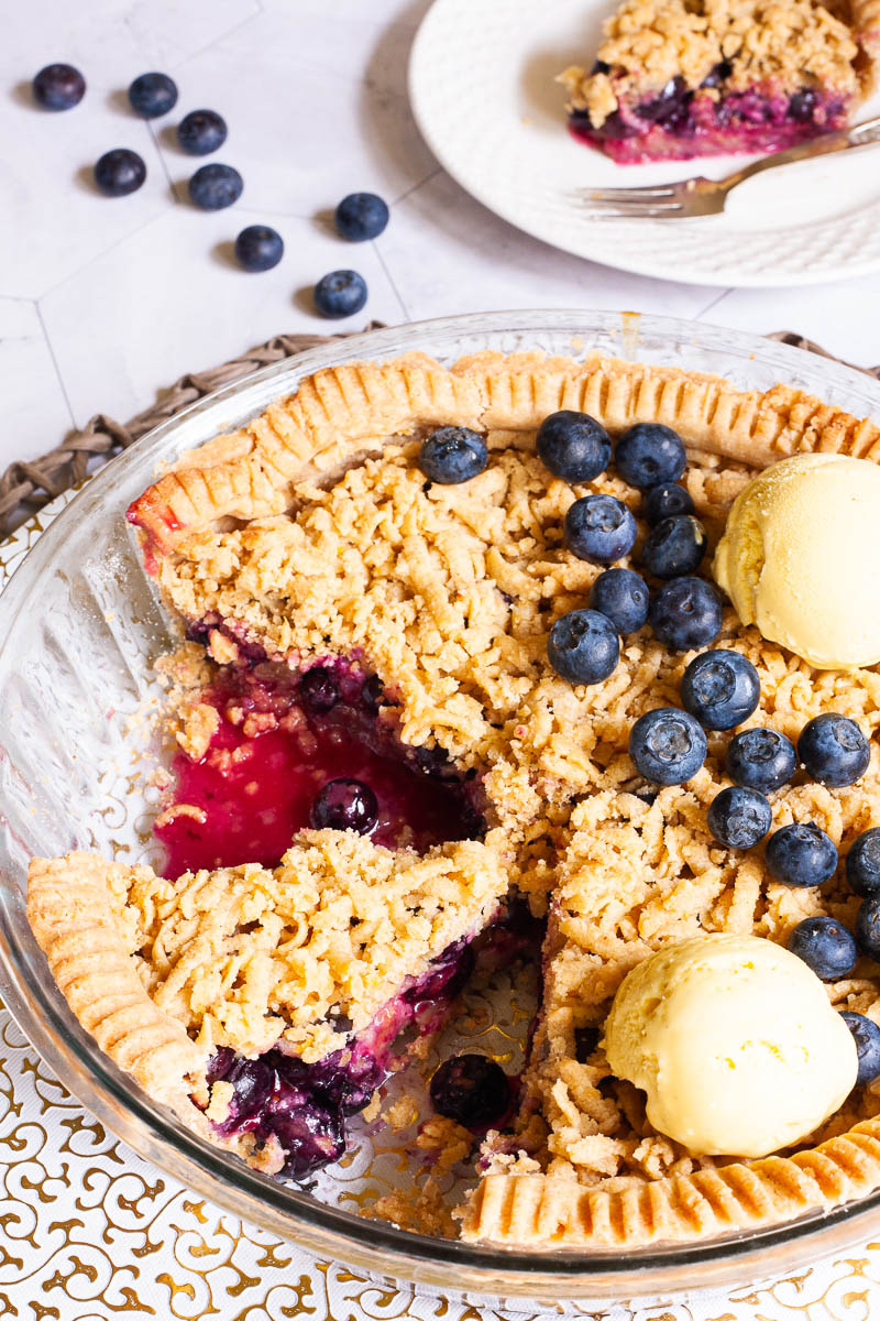 A whole blueberry pie from above with fresh blueberries on top as decoration, crumble topping and 2 scoops of yellow ice cream. 2 slices are missing. A plate with one slice is in the background. Fresh blueberries are scattered around.