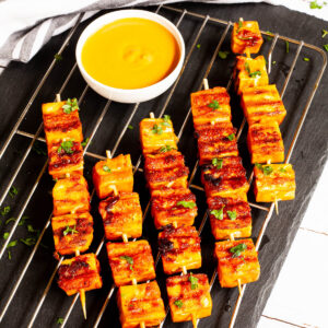 Yellow-brown tofu cubes on wooden skewers are criss cross over each other on a grilling rack placed on a black board. A small white bowl is next to them with a yellow dipping sauce.