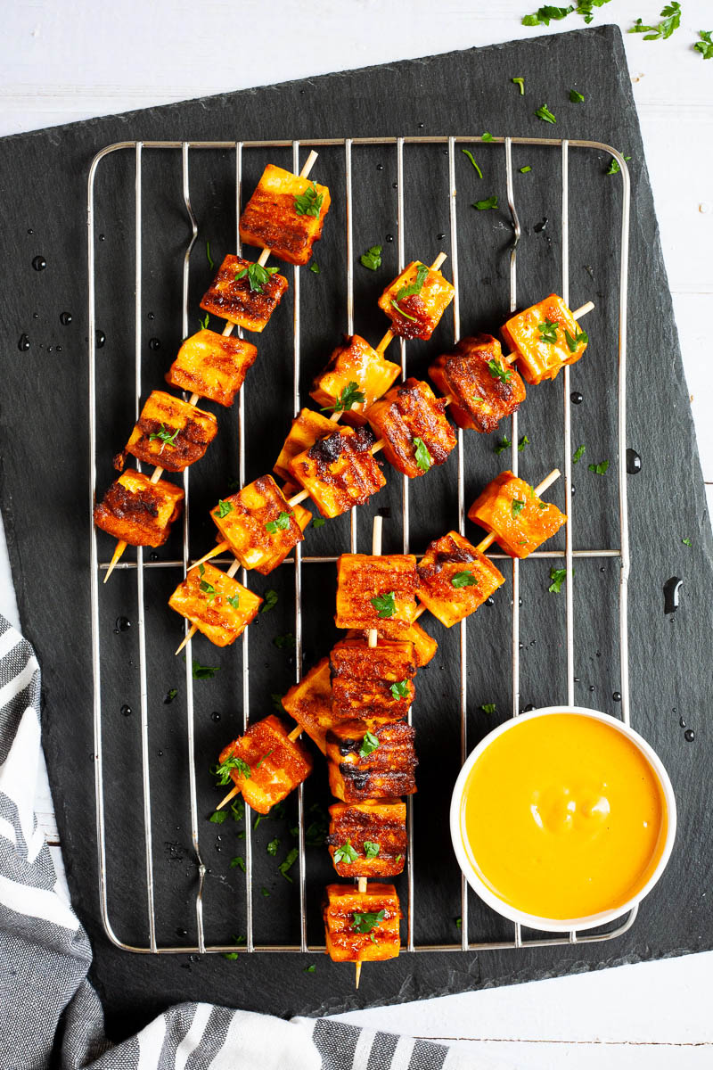 Yellow-brown tofu cubes on wooden skewers are criss cross over each other on a grilling rack placed on a black board. A small white bowl is next to them with a yellow dipping sauce.