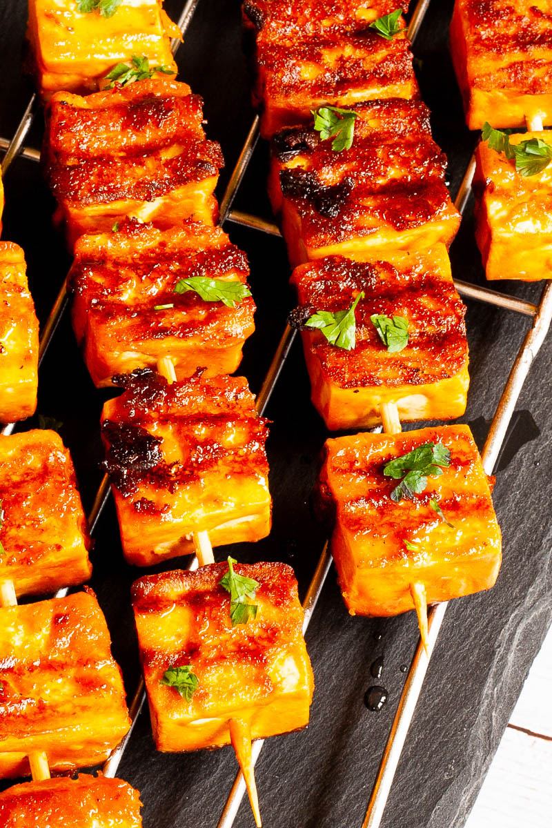 Yellow-brown tofu cubes on wooden skewers are criss cross over each other on a grilling rack placed on a black board.