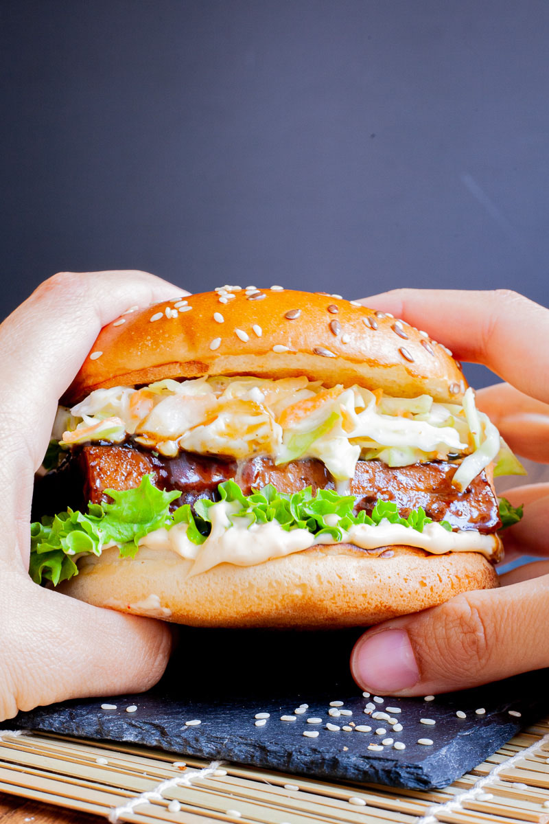 Two hands are holding a burger bun up with white sauce, green ruffled lettuce, dark glazed tofu slice, shredded cabbage and carrot in white sauce layers.