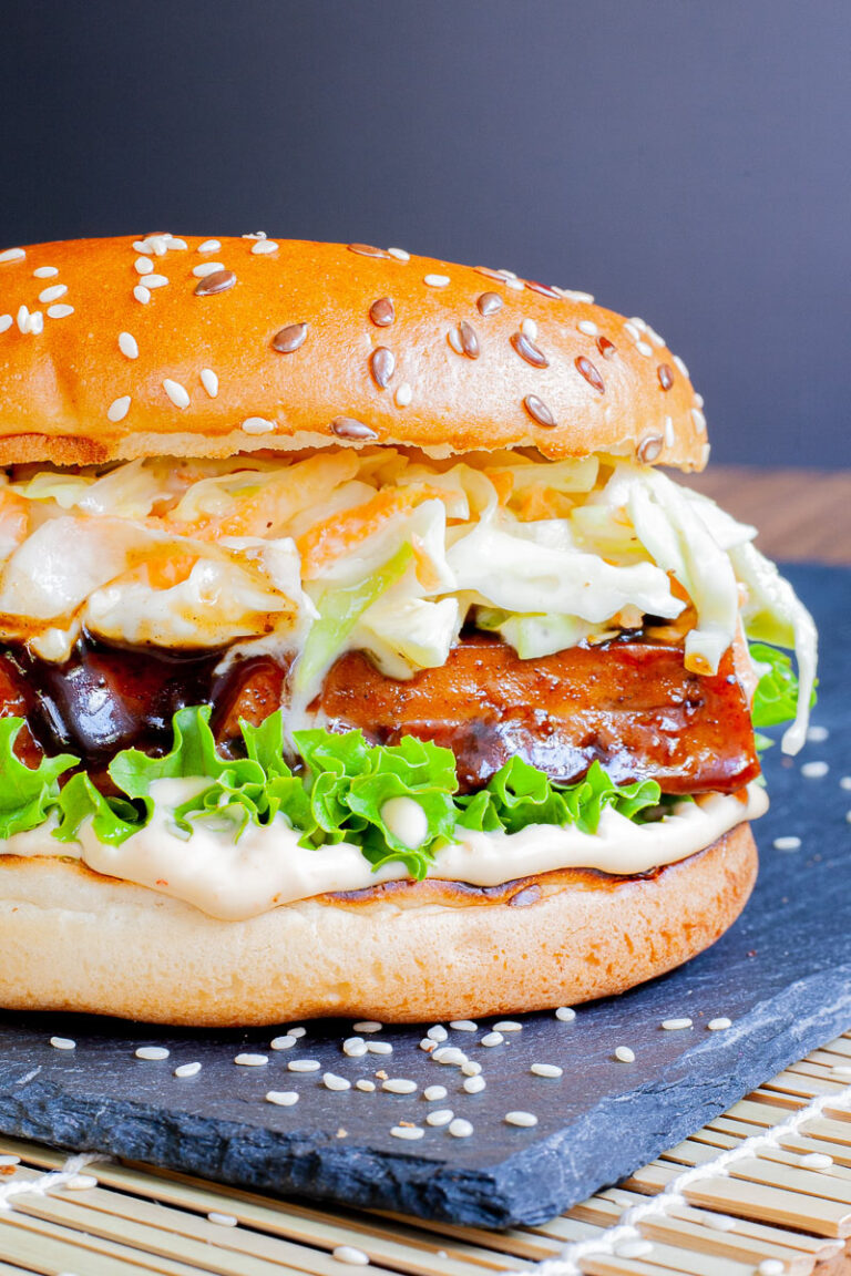 A burger bun up close with white sauce, green ruffled lettuce, dark glazed tofu slice, shredded cabbage and carrot in white sauce.