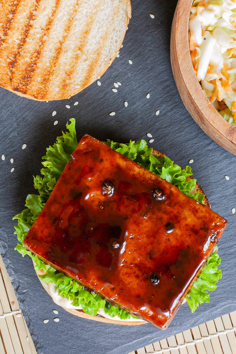A burger from above with green ruffled lettuce and a dark glazed tofu slice on top. The top bun is next to it with charcoal marks. A small woddon bowl is in the corner with shredded carrot and cabbage in white sauce.