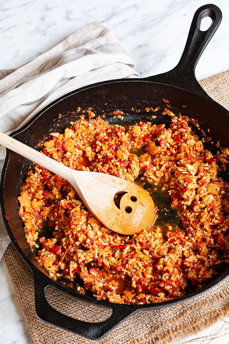 A black cast iron skillet from above with yellow brown pieces in a red chunky sauce. There is a wooden spatula with a smiley face is stirring the middle.