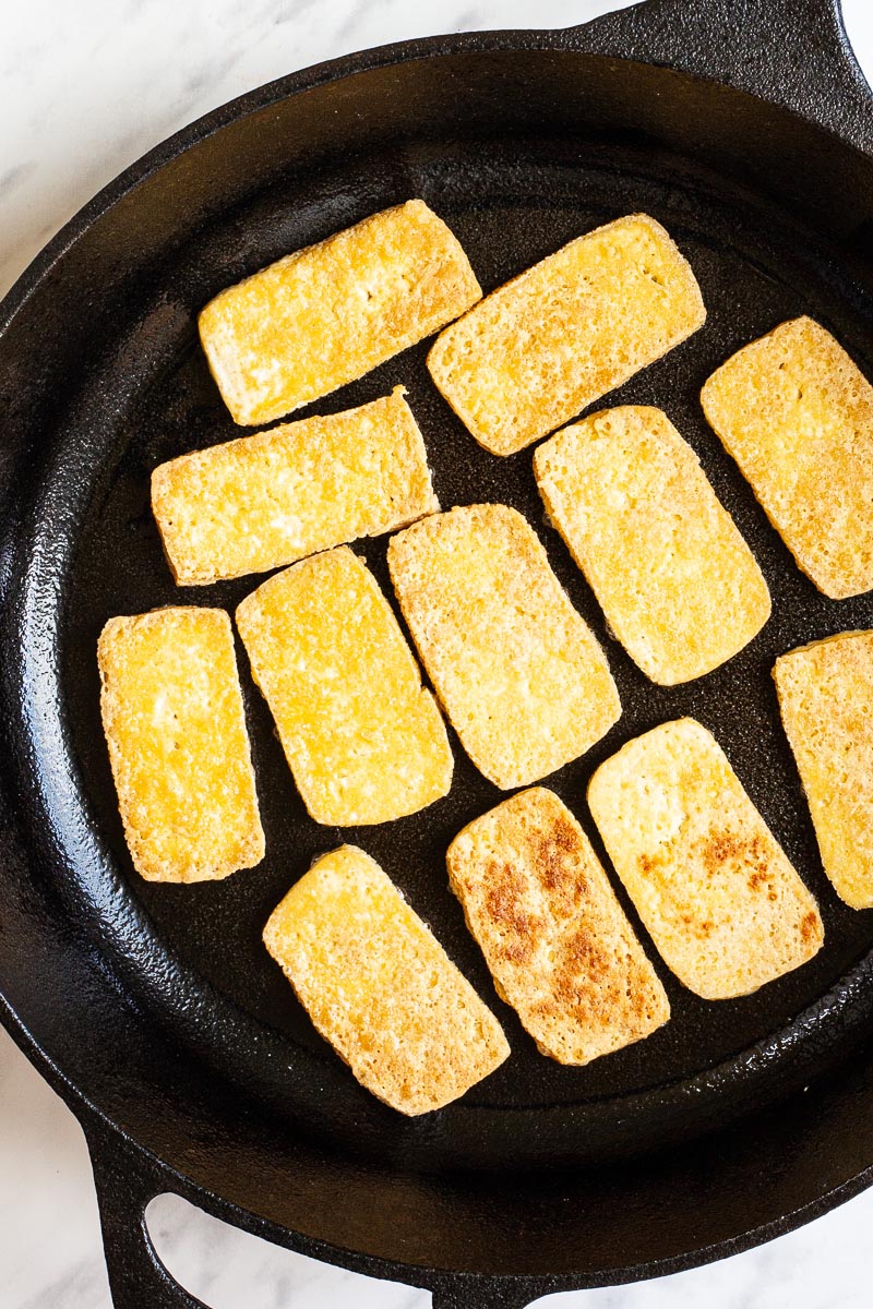 A black cast iron skillet from above with yellow tofu slices fried until golden brown and crispy