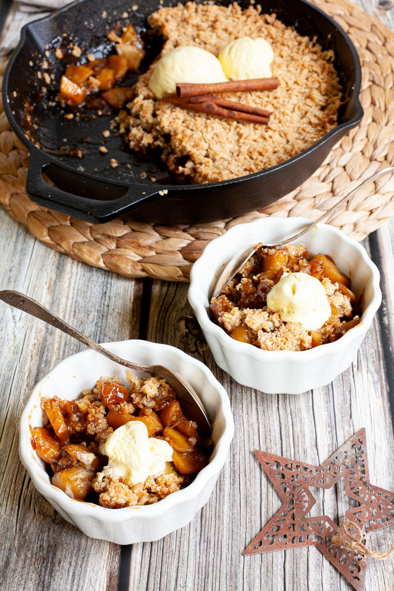 2 small white bowls with diced apples in caramel sauce, crumble topping and a scoop of yellow ice cream. A black skillet is in the background with the remaining crumbles.