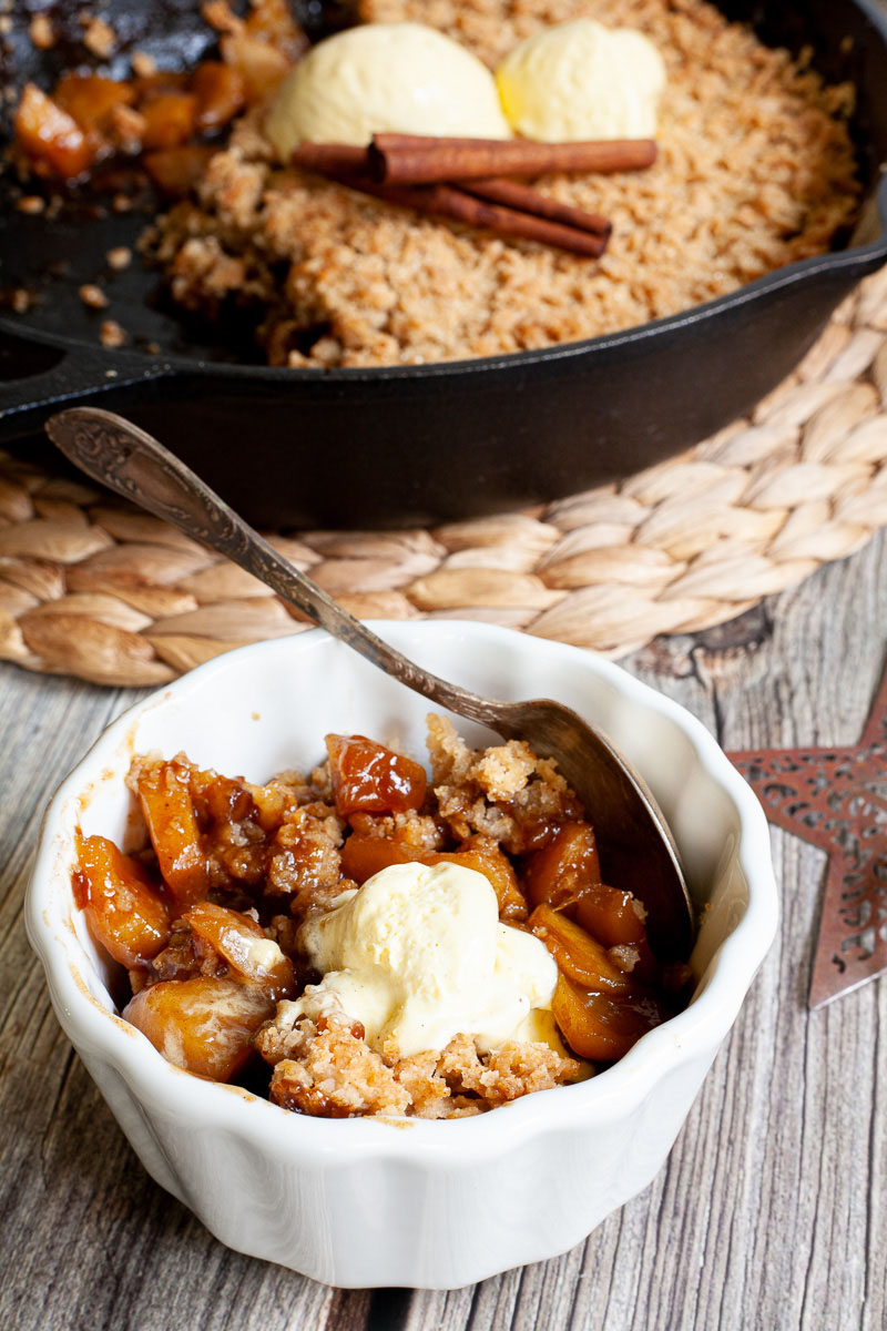 Small white bowl with diced apples in caramel sauce, crumble topping and a scoop of yellow ice cream. A black skillet is in the background with the remaining crumbles.