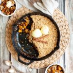 A black skillet from above with diced apples in caramel sauce, crumble topping and 2 scoops of yellow ice cream. 2 small whie bowls is filled with the apple crumble hence there is a part missing in the skillet.
