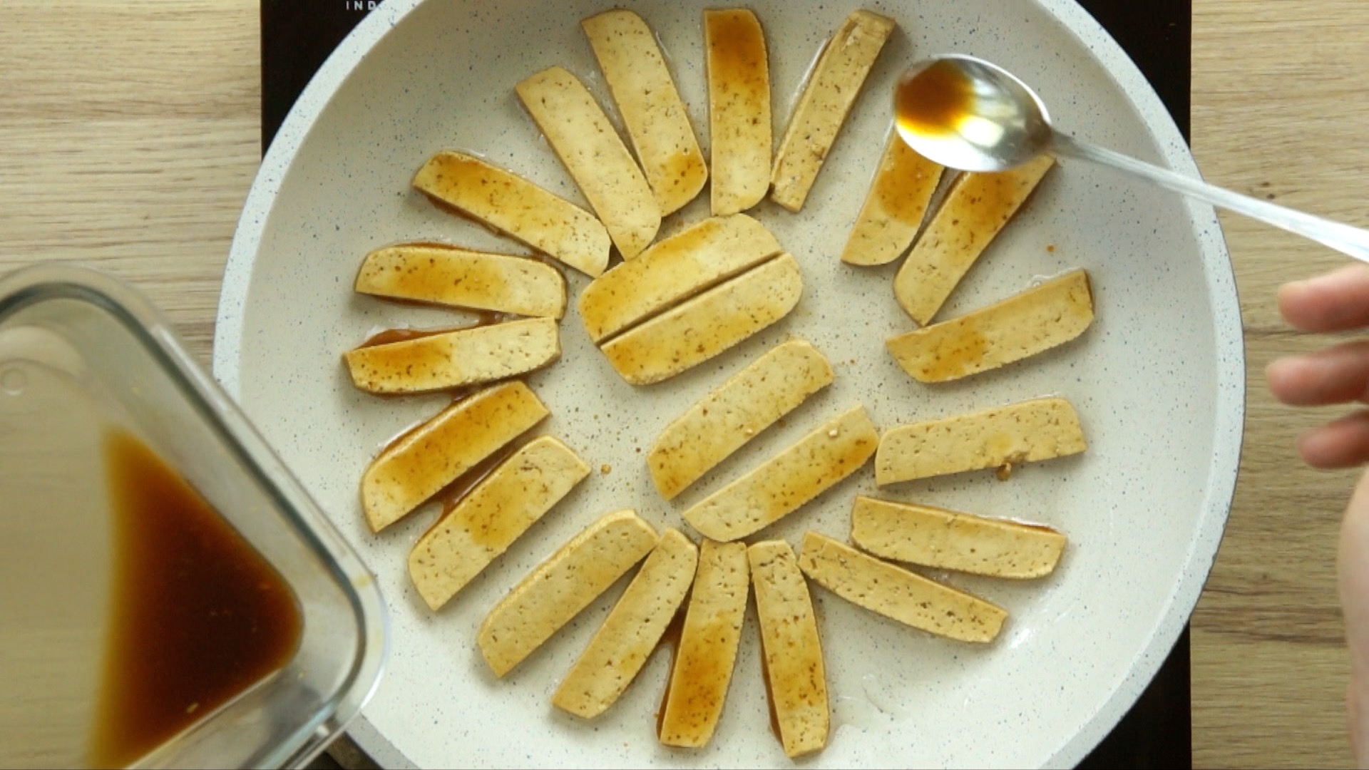 A white frying pan with light brown tofu slices and a spoon with dark brown sauce.