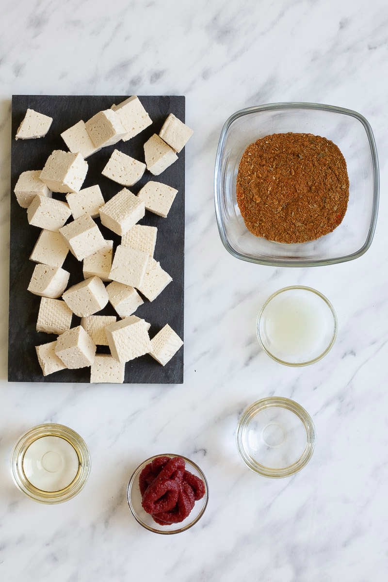 Ingredients of red curry with tofu in small glass bowls translucent liquids, red paste and red spice blend. White tofu cubes are displayed on a black plate.