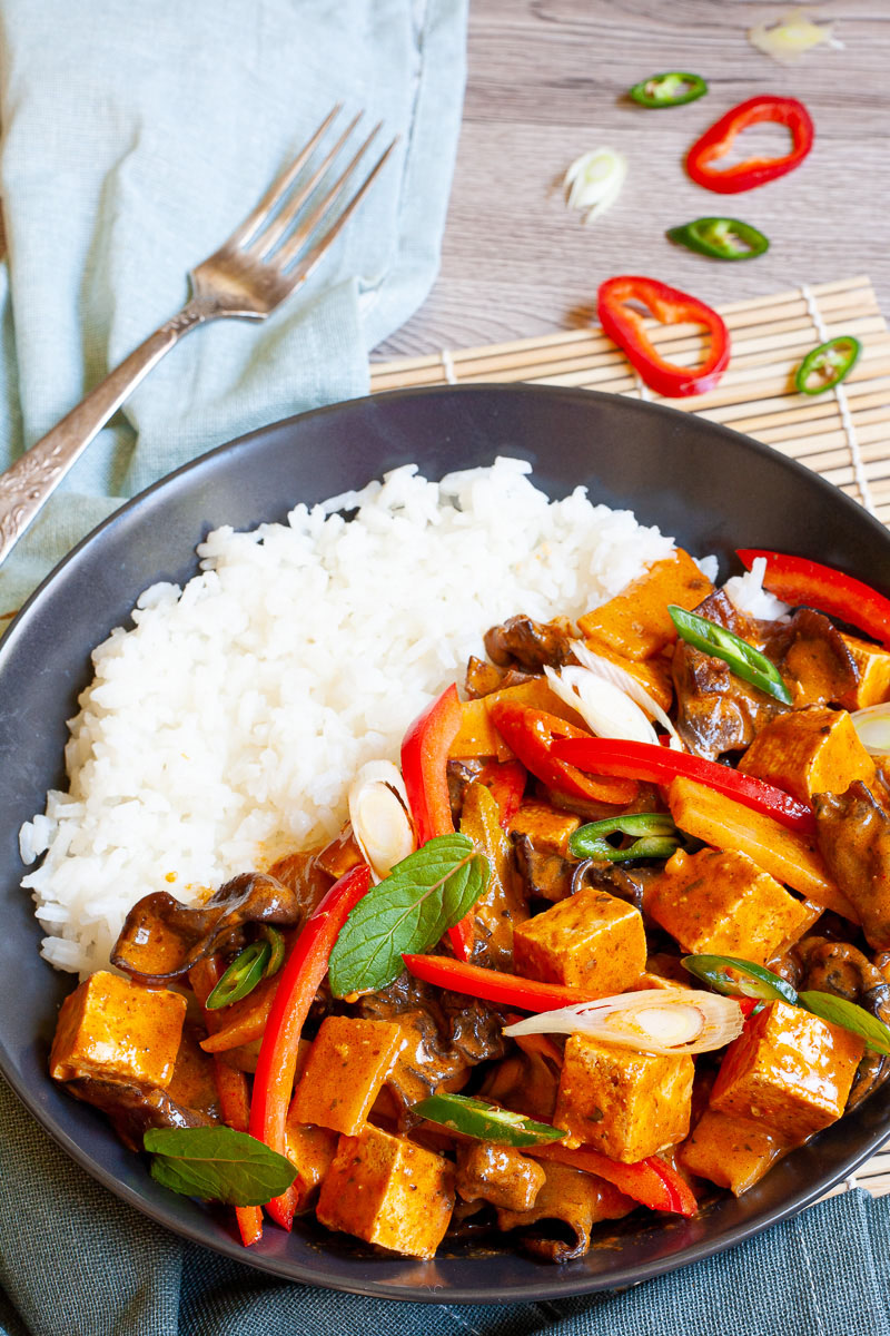 A black bowl of rice and brown red sauce with tofu cubes, sliced bell peppers, sliced green chilies, mint leaves, sliced spring onion, mushrooms slices.