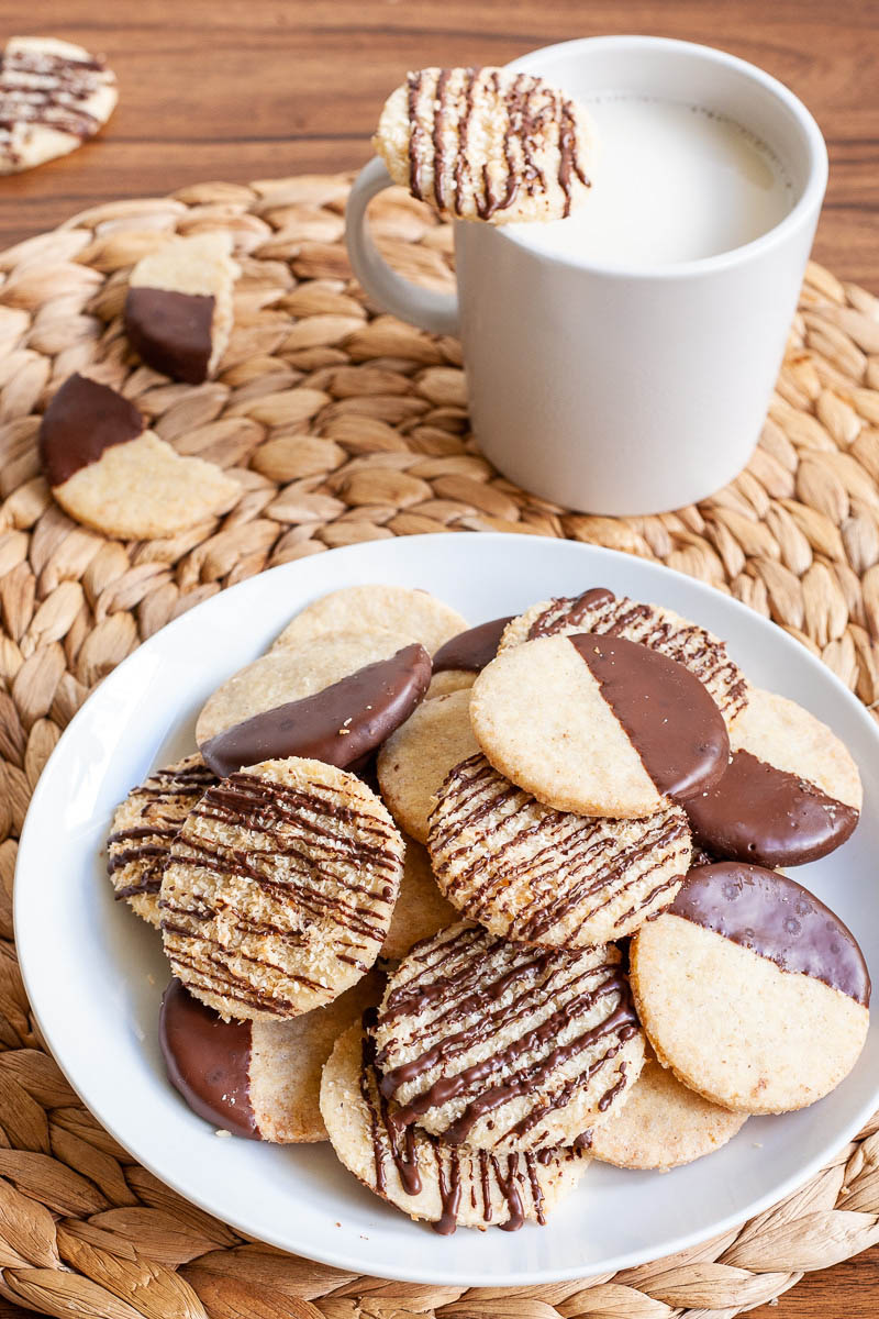 A white plate is full of thin round cookies placed on top of each other. Some cookies are partly covered in chocolate, some cookies are drizzled with chocolate so brown stripes can be seen. There is a mug of milk in the background with broken cookies around.