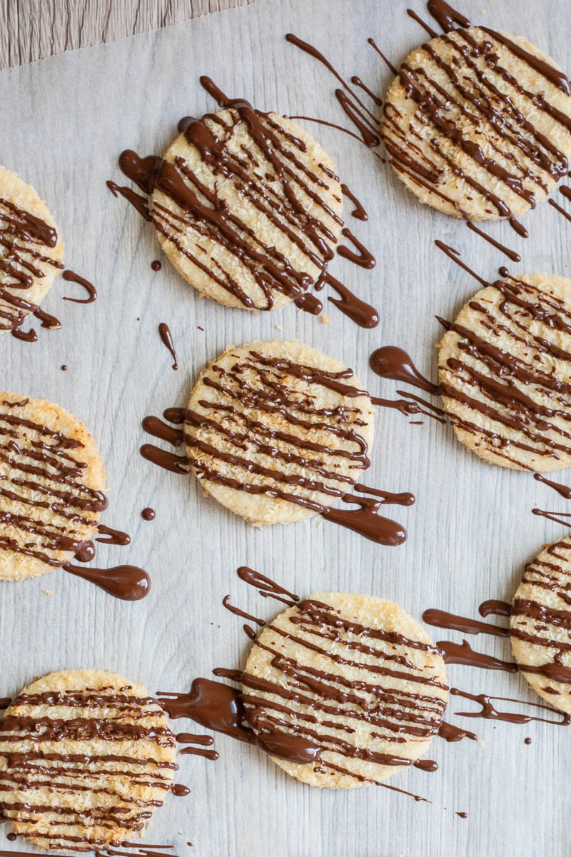 Several thin round cookies on a parchment paper. Brown stripes of melted chocolate is on top as they were just drizzled with it.